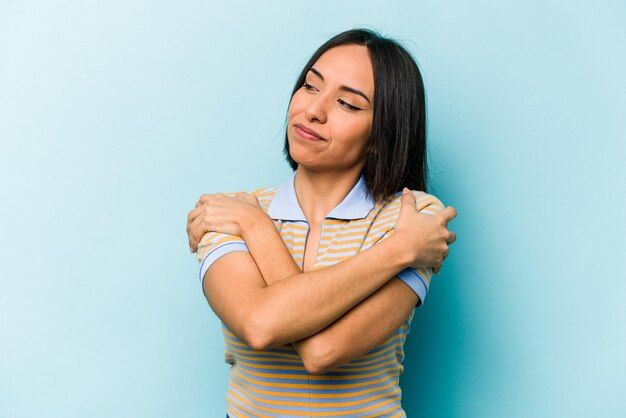 Joven hispana aislada de fondo azul abraza sonriendo despreocupada y feliz