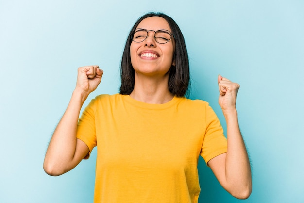 Joven hispana aislada de fondo azul celebrando una victoria, pasión y entusiasmo, expresión feliz