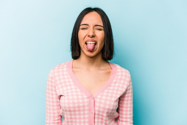 Joven hispana aislada de fondo azul divertida y amigable sacando la lengua