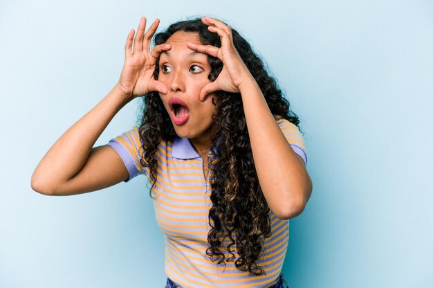 Joven hispana aislada de fondo azul manteniendo los ojos abiertos para encontrar una oportunidad de éxito