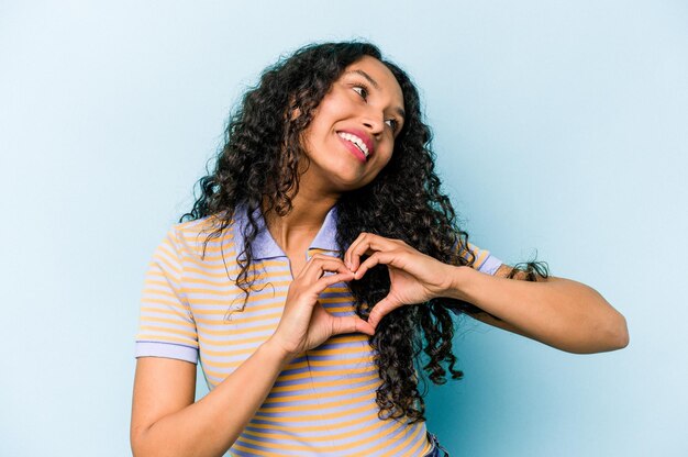 Joven hispana aislada de fondo azul sonriendo y mostrando una forma de corazón con las manos