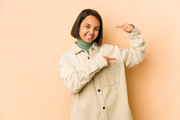 Joven hispana aislada sosteniendo algo pequeño con los dedos índices, sonriente y confiada.