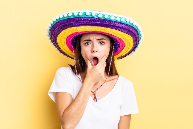 Joven hispana con la boca y los ojos bien abiertos y la mano en la barbilla. concepto de sombrero mexicano