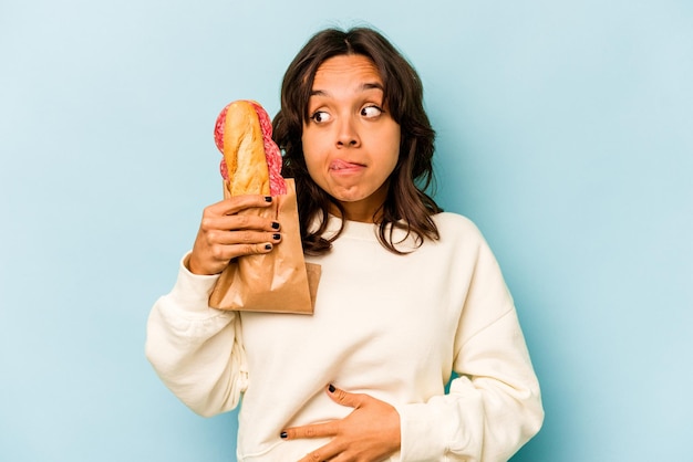 Joven hispana comiendo un sándwich aislado sobre fondo azul.