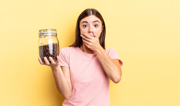 Joven hispana cubriendo la boca con las manos con una sorpresa. concepto de granos de cafe