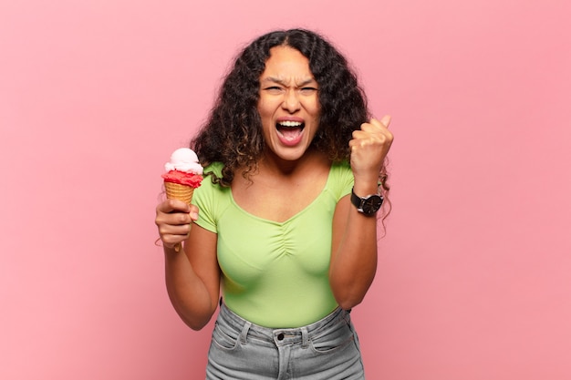 Joven hispana gritando agresivamente con una expresión de enojo o con los puños cerrados celebrando el éxito. concepto de helado