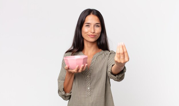 Joven hispana haciendo gesto de capice o dinero, diciéndole que pague y sosteniendo un recipiente o una olla vacía