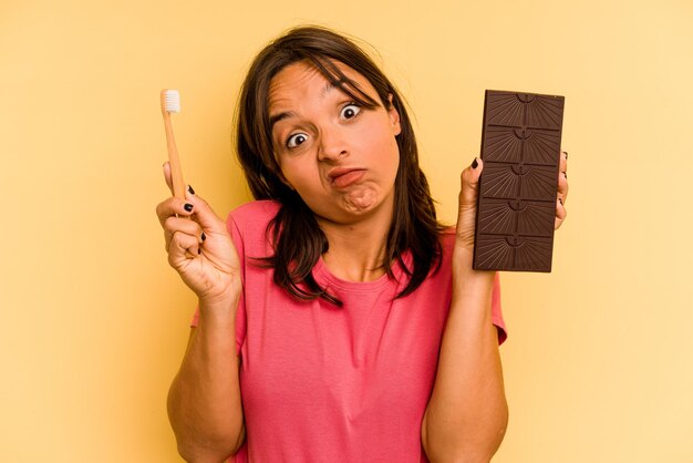 Joven hispana lavándose los dientes después de comer chocolate aislado de fondo amarillo