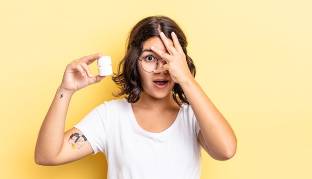 Joven hispana mirando conmocionado, asustado o aterrorizado, cubriéndose el rostro con la mano. concepto de píldoras de enfermedad