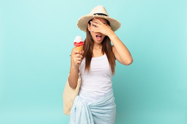 Joven hispana mirando conmocionado, asustado o aterrorizado, cubriéndose el rostro con la mano y sosteniendo un helado. concepto sumergible