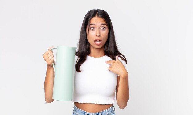 Joven hispana mirando consternado y sorprendido con la boca abierta, apuntando a sí mismo y sosteniendo un termo de café