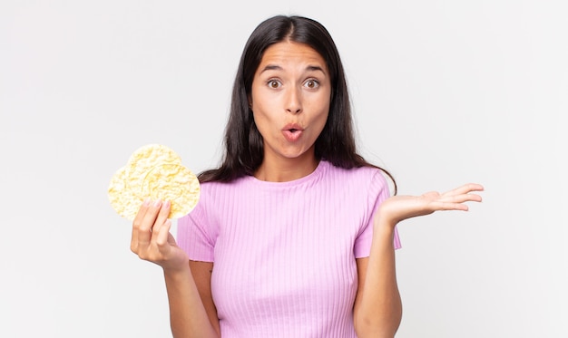 Joven hispana mirando sorprendida y consternada, con la mandíbula caída sosteniendo un objeto y sosteniendo una galleta de arroz. concepto de dieta