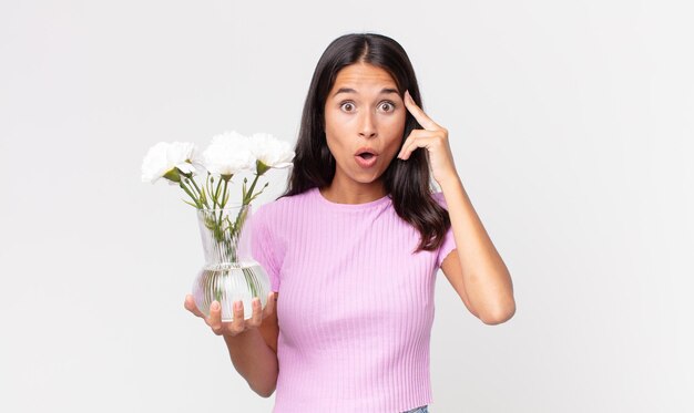 Joven hispana mirando sorprendido, dándose cuenta de un nuevo pensamiento, idea o concepto sosteniendo flores decorativas