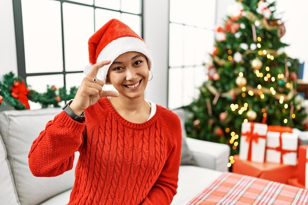 Joven hispana con el pelo corto y sombrero de navidad sentada en el sofá sonriendo y confiada gesticulando con la mano haciendo un signo de tamaño pequeño con los dedos mirando y el concepto de medida de la cámara