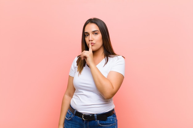 Joven hispana pidiendo silencio y silencio, haciendo un gesto con el dedo frente a la boca, diciendo shh o guardando un secreto en la pared rosa