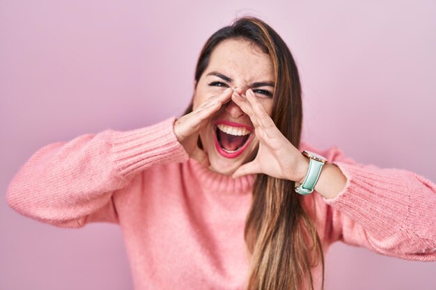 Joven hispana de pie sobre fondo rosa gritando enojada con las manos sobre la boca