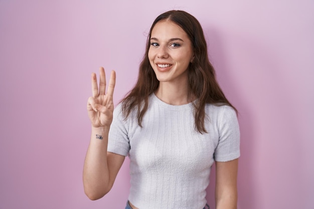 Joven hispana de pie sobre fondo rosa mostrando y señalando con los dedos número tres mientras sonríe confiada y feliz