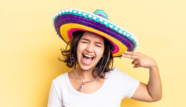 Joven hispana que parece infeliz y estresada, gesto suicida haciendo señas de armas. concepto de sombrero mexicano