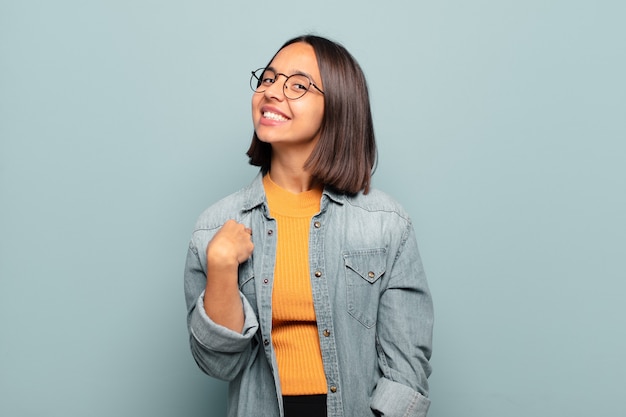 Joven hispana que parece orgullosa, segura y feliz, sonriendo y apuntando a sí misma o haciendo la señal número uno