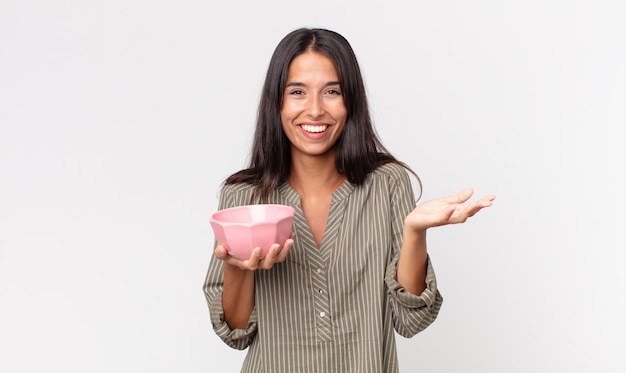 Joven hispana que se siente feliz, sorprendida al darse cuenta de una solución o idea y sosteniendo un recipiente o una olla vacía