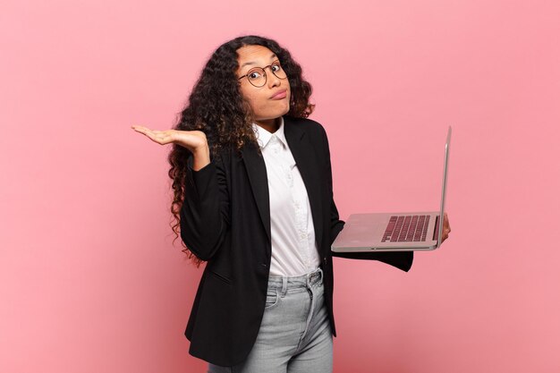 Joven hispana que se siente perpleja y confundida, dudando, ponderando o eligiendo diferentes opciones con expresión divertida. concepto de laptop