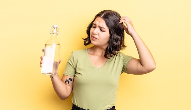 Joven hispana que se siente perpleja y confundida, rascándose la cabeza. concepto de botella de agua