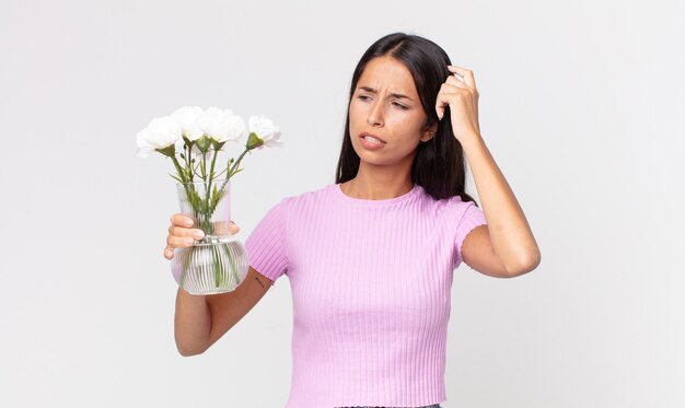 Joven hispana que se siente perpleja y confundida, rascándose la cabeza sosteniendo flores decorativas