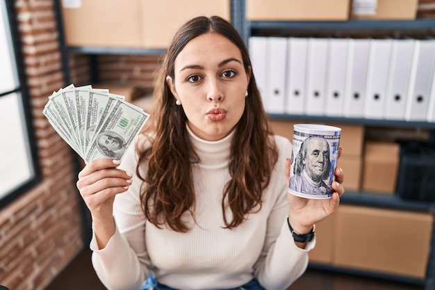 Joven hispana que trabaja en el comercio electrónico de pequeñas empresas con dinero y una alcancía mirando a la cámara lanzando un beso siendo encantadora y sexy. expresión de amor.