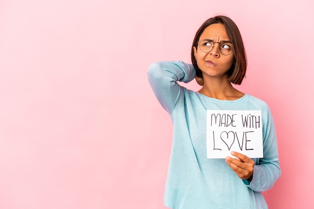 Joven hispana de raza mixta sosteniendo un cartel de papel hecho con amor tocando la parte posterior de la cabeza, pensando y tomando una decisión.