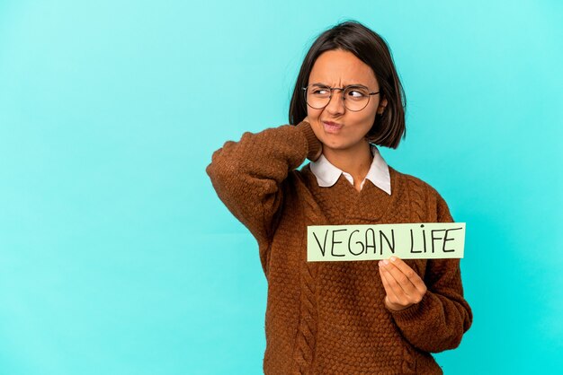 Foto joven hispana de raza mixta sosteniendo un cartel de vida vegana tocando la parte posterior de la cabeza, pensando y tomando una decisión.