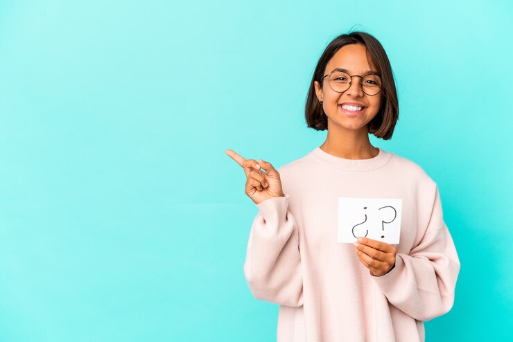 Foto joven hispana de raza mixta sosteniendo un tablero de interrogación sonriendo y apuntando a un lado, mostrando algo en el espacio en blanco.