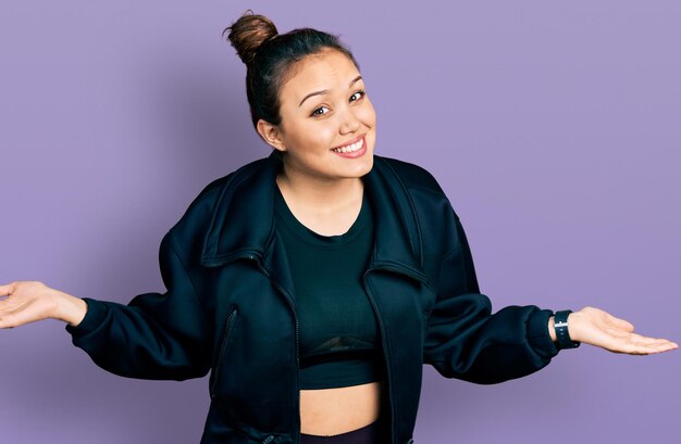 Joven hispana con ropa deportiva sonriendo mostrando ambas manos palmas abiertas, presentando y publicitando comparación y equilibrio