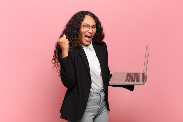 Joven hispana sintiéndose conmocionada, emocionada y feliz, riendo y celebrando el éxito, diciendo ¡guau !. concepto de laptop