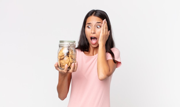 Joven hispana sintiéndose feliz, emocionada y sorprendida y sosteniendo una botella de vidrio de galletas