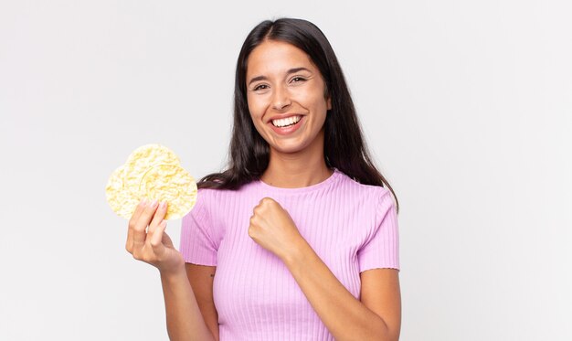 Joven hispana sintiéndose feliz y enfrentando un desafío o celebrando y sosteniendo una galleta de arroz. concepto de dieta