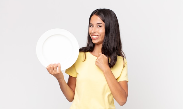 Foto joven hispana sintiéndose feliz y enfrentando un desafío o celebrando y sosteniendo un plato vacío