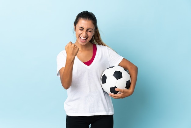 Joven hispana sobre pared azul aislada con balón de fútbol celebrando una victoria