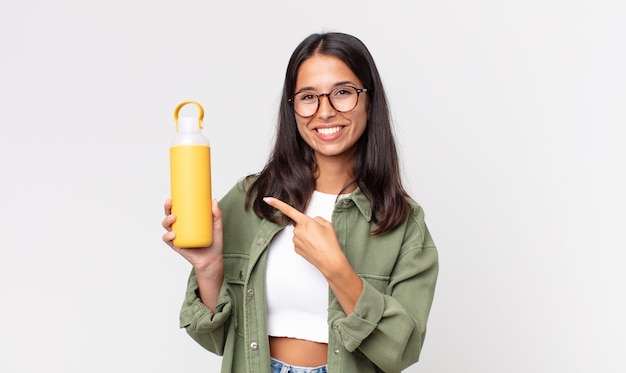 Joven hispana sonriendo alegremente, sintiéndose feliz y apuntando hacia un lado y sosteniendo un termo de café