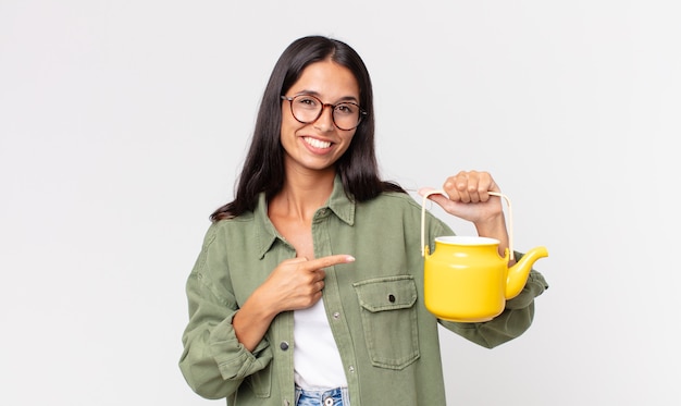 Joven hispana sonriendo alegremente, sintiéndose feliz y apuntando hacia un lado y sosteniendo una tetera