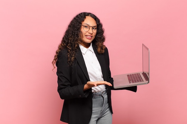 Joven hispana sonriendo alegremente, sintiéndose feliz y mostrando un concepto en el espacio de la copia con la palma de la mano. concepto de laptop