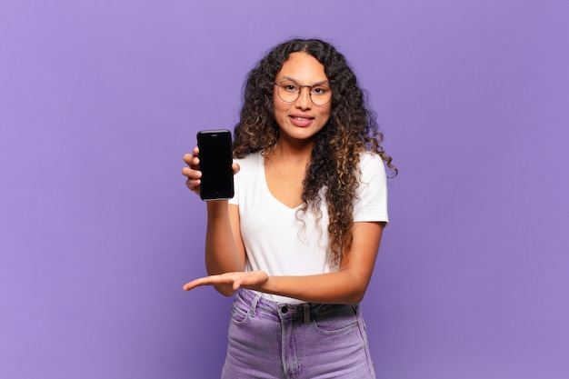 Joven hispana sonriendo alegremente, sintiéndose feliz y mostrando un concepto en el espacio de la copia con la palma de la mano. concepto de teléfono inteligente