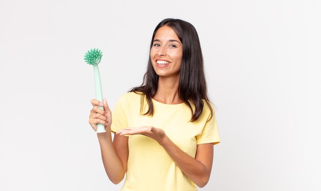 Joven hispana sonriendo alegremente, sintiéndose feliz y mostrando un concepto y sosteniendo un cepillo de limpieza de platos