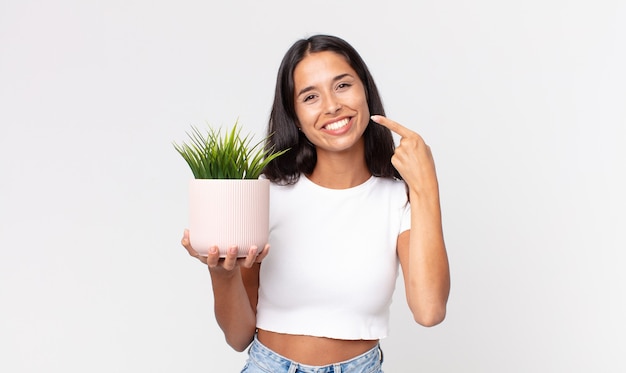 Joven hispana sonriendo con confianza apuntando a su propia amplia sonrisa y sosteniendo una planta decorativa de la casa