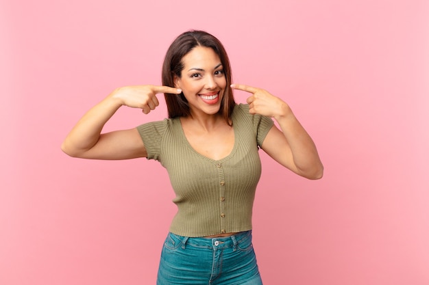 Joven hispana sonriendo con confianza apuntando a su propia amplia sonrisa