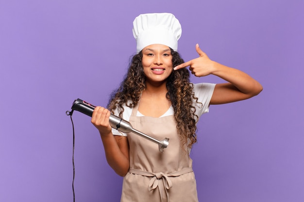 Joven hispana sonriendo con confianza apuntando a su propia sonrisa amplia, actitud positiva, relajada y satisfecha. concepto de chef