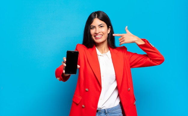 Joven hispana sonriendo con confianza apuntando a su propia sonrisa amplia, actitud positiva, relajada y satisfecha. espacio de copia de la pantalla del teléfono