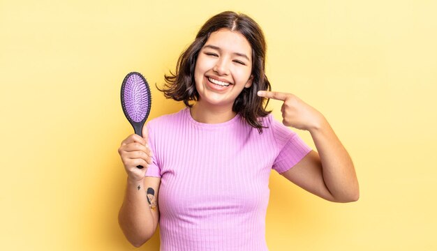 joven hispana sonriendo con confianza apuntando a su propia sonrisa amplia. concepto de cepillo para el cabello