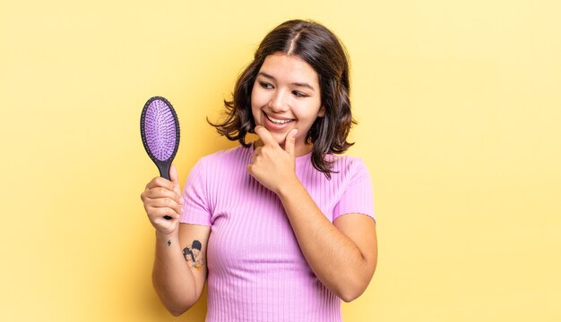 Joven hispana sonriendo con una expresión feliz y segura con la mano en la barbilla. concepto de cepillo para el cabello