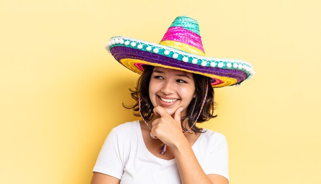 Joven hispana sonriendo con una expresión feliz y segura con la mano en la barbilla. concepto de sombrero mexicano