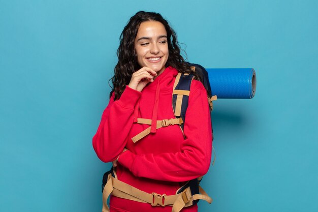 Joven hispana sonriendo con una expresión feliz y segura con la mano en la barbilla, preguntándose y mirando hacia un lado